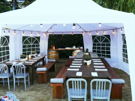 A white outdoor tent with string lights, wooden tables, gray chairs, benches, placeholders, candles, and barrels inside, set up for an event.