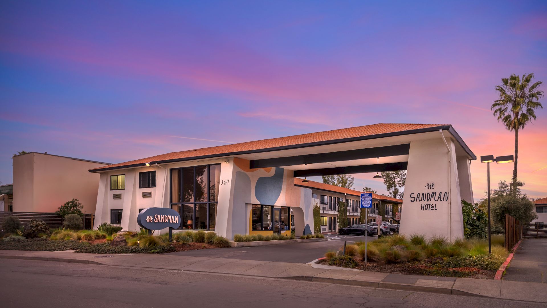 The image shows a mid-century modern hotel exterior at dusk with a sign and distinctive architectural features, surrounded by landscaping.