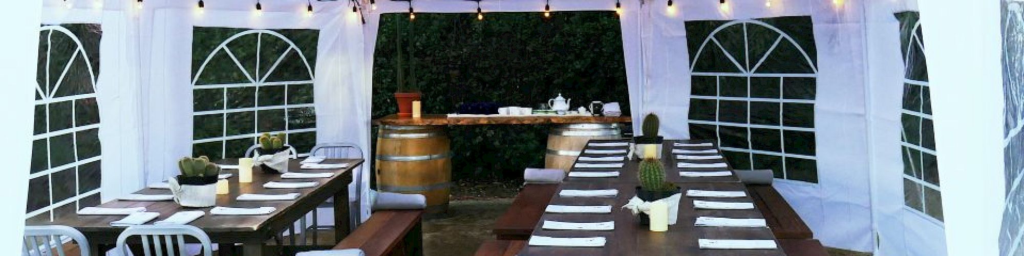 Outdoor setup with long wooden tables and benches under a white canopy, decorated with string lights, ready for a gathering or meal.