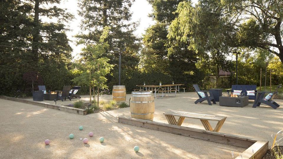 The image shows an outdoor space with seating and game setups, surrounded by tall trees. There are game balls scattered on the ground.