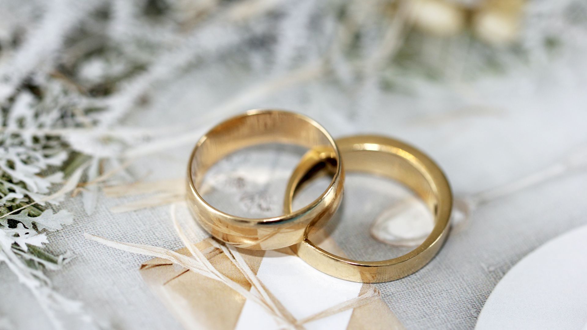 Two gold wedding rings are placed on a light-colored surface with delicate, dried foliage and a white envelope.