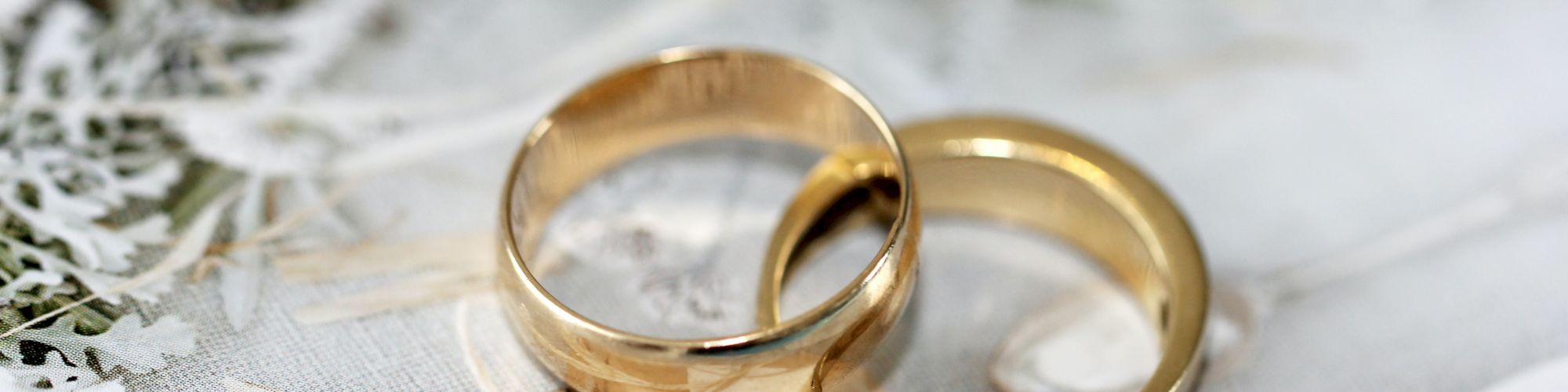 Two gold wedding rings are placed on a light-colored surface with delicate, dried foliage and a white envelope.