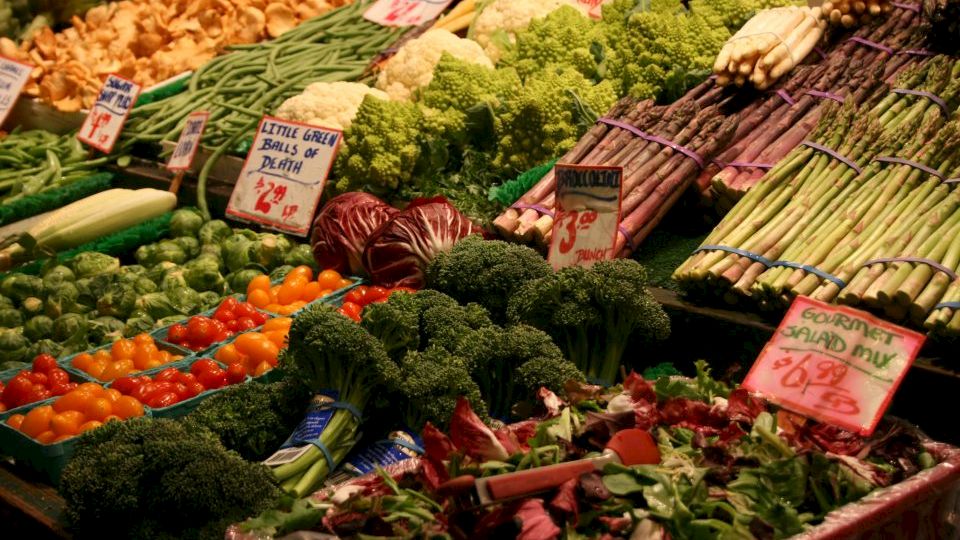 The image shows a vibrant market stand with a variety of fresh vegetables, including asparagus, tomatoes, broccoli, and leafy greens, labeled with prices.
