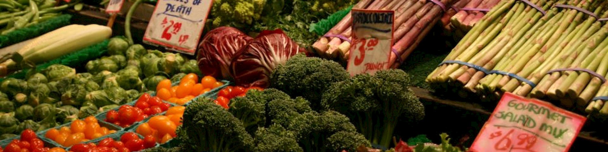 The image shows a vibrant market stand with a variety of fresh vegetables, including asparagus, tomatoes, broccoli, and leafy greens, labeled with prices.