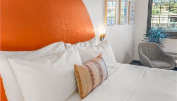 A neatly made bed with white linens and colorful pillows, adjacent to a gray armchair, with wall art and a window in the background ending the sentence.