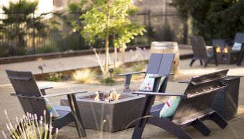 The image shows an outdoor seating area with black chairs arranged around a fire pit. There are cushions on the chairs, and plants surround the area.