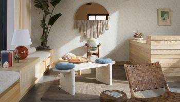 A cozy room with modern furniture, a woven chair, a round mirror, and plants. Decor includes books, a globe, and a table with cushions.