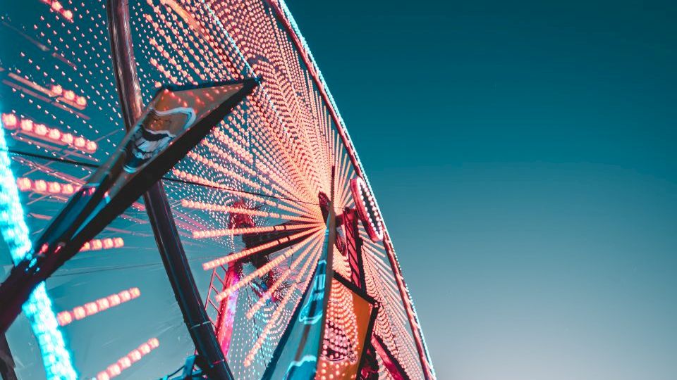 The image shows a close-up of a large Ferris wheel, brightly illuminated with colorful lights against a clear, deep blue sky.