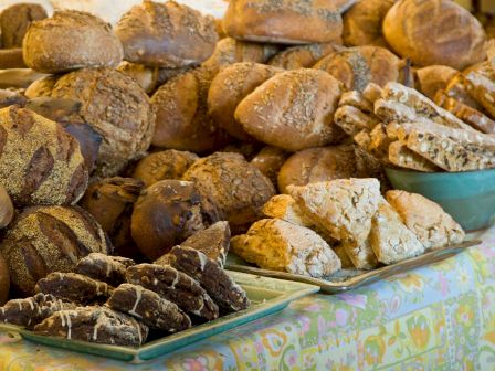 The image shows a variety of bread and pastries displayed on a table with patterned cloth. There are loaves, scones, and pastries in the scene.