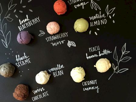 This image shows various flavors of ice cream scoops arranged on a black surface, each labeled: LOCAL BLACKBERRY, STRAWBERRY MISO, MATCHA ALMOND, BLACK SESAME, PEACH BROWN BUTTER, VANILLA BEAN, COCONUT CURRY, and SWISS CHOCOLATE.