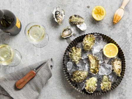 A plate of oysters on ice, two glasses of white wine, a bottle, a knife, half a lemon, a citrus juicer, and a napkin on a textured surface.