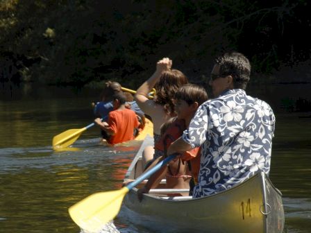 People are canoeing on a calm body of water, with three canoes visible in a single file line, paddling under the sun.