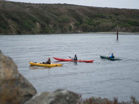 Three people are kayaking on a body of water with a hilly, tree-lined shore in the background.