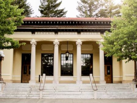 The image shows a classical building with columns, steps, and trees in front, bathed in sunlight. It has large windows and an ornate facade.