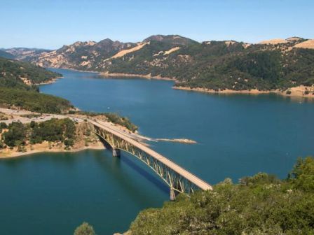 A scenic view of a bridge spanning over a large body of water surrounded by tree-covered hills and distant mountains under a clear blue sky.