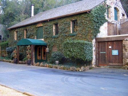 The image depicts an ivy-covered stone building with green trims, a wooden door, and a small awning over the entrance, surrounded by greenery.