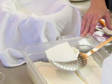 A person's hand is using a slotted spoon to lift a piece of white curd or tofu from a container with a cloth-lined bowl beside it.