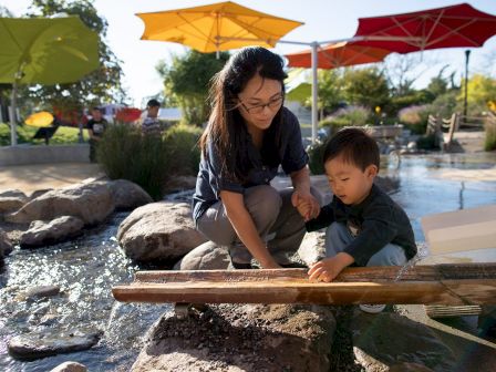 A woman and a child are playing by a shallow stream with rocks, under colorful umbrellas, on a sunny day.