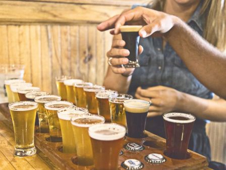 The image shows several glasses of beer, some dark and some light, with a person placing a glass on a wooden table.