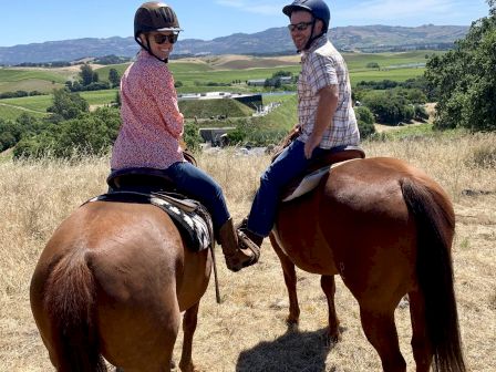 Two people wearing helmets are riding horses on a scenic countryside trail with rolling hills and a building in the background.