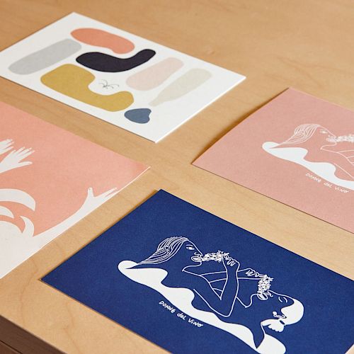 The image shows four colorful art prints on a wooden surface, featuring abstract and figurative designs in various colors and styles.