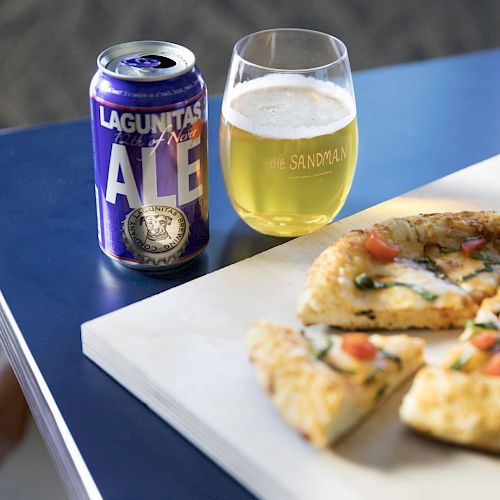 A can of Lagunitas Ale, a glass of beer, and a pizza with toppings on a cutting board are on a table.