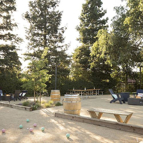 The image shows an outdoor recreational area with seating, tables, barrels, and bocce balls, surrounded by tall trees and greenery.