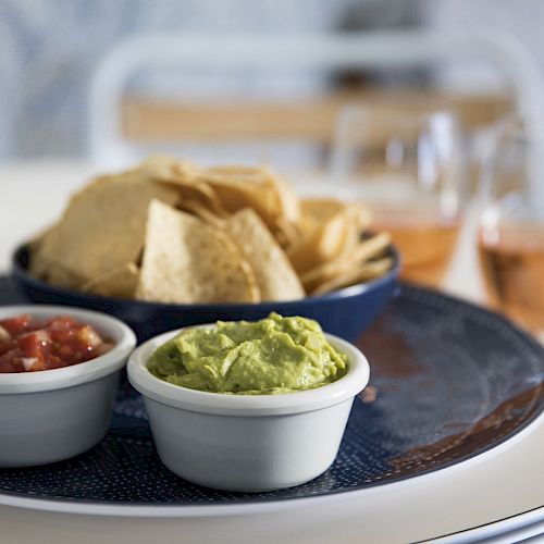 A plate with tortilla chips, salsa, and guacamole in small bowls; two glasses of pink-colored drink are in the background.