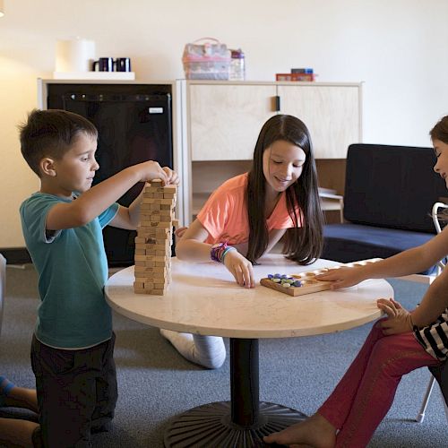 Three children are playing games at a round table in a cozy room, with a lamp, shelves, and books in the background.