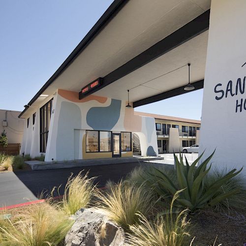 The image shows the exterior of The Sandman Hotel, featuring a modern design and its signage, with landscaping in the foreground.