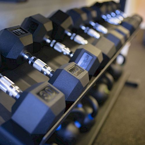 The image shows a rack of hexagonal dumbbells of various weights in a gym setting with a blurred background.