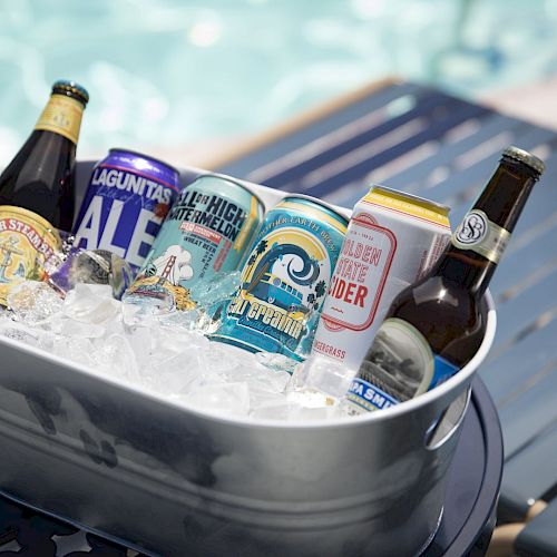 A metal bucket filled with ice holds various bottles and cans of beer, placed on a table beside a swimming pool.