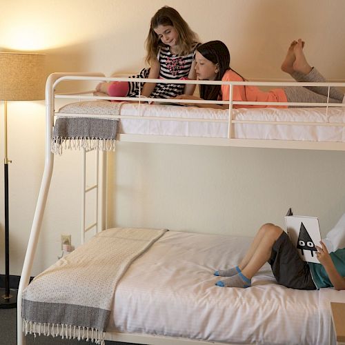 Three children are in a room with bunk beds; two are on the top bunk, and one is reading a book on the bottom bunk.