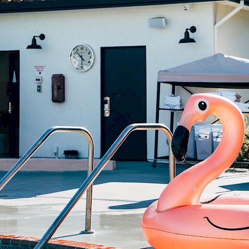 An inflatable pink flamingo float in a swimming pool, with a poolside building and a clock in the background, surrounded by trees.