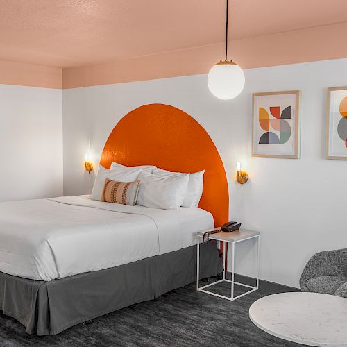 The image shows a modern bedroom with a queen bed, abstract art, an orange accent wall, a round pendant light, a side table, a cozy chair, and a small table.