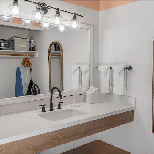 A modern bathroom with a vanity mirror, sink, black faucet, and towels, featuring a closet with a safe, hangers, and an iron in the background.