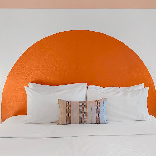 A neatly made bed with white bedding, centered below an orange semicircle wall design, flanked by two wall-mounted lights, and a framed artwork.