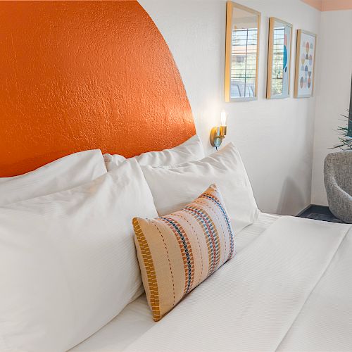 A cozy bedroom with a white bed, orange accent wall, and decorative pillows. There is a gray chair by the window with a plant and framed pictures.