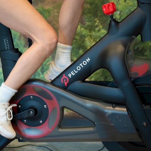 A person is using a Peloton stationary bike, showing their legs pedaling while wearing white socks and shoes, against a backdrop of greenery.