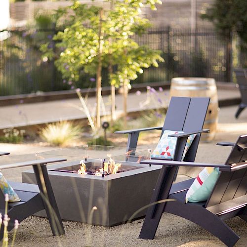 The image shows an outdoor patio area with four Adirondack chairs around a square fire pit, surrounded by plants and additional seating in the background.