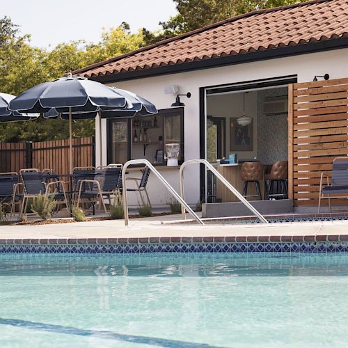 The image shows a pool with blue water, surrounded by a deck with chairs and umbrellas, next to a building with a tiled roof.