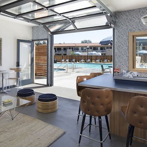 This image shows a modern, open-concept room with a bar area, stools, a glass coffee table, and a view of an outdoor pool.