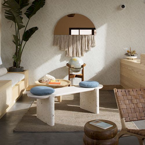 A cozy, modern living room with a mix of natural textures and decor, featuring a mirror, bench seating, woven chair, plants, and a small coffee table.