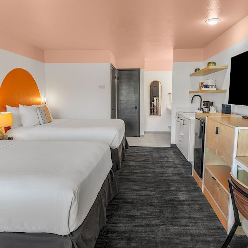 A modern hotel room with two beds, a TV, a desk, a mirror, and a kitchenette, featuring orange and white decor and stylish furnishings.