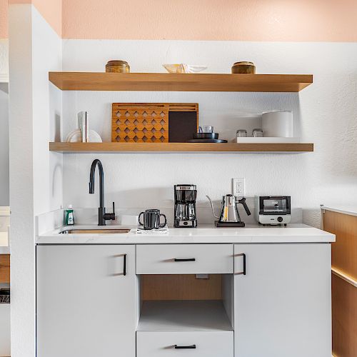 This image shows a kitchen area with open shelves, a sink, coffee maker, toaster, microwave, and a mini fridge. A TV is mounted on the wall.