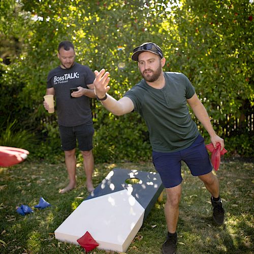 Two men are playing cornhole outdoors. One is throwing a bean bag, and the other watches. Greenery and trees are in the background.