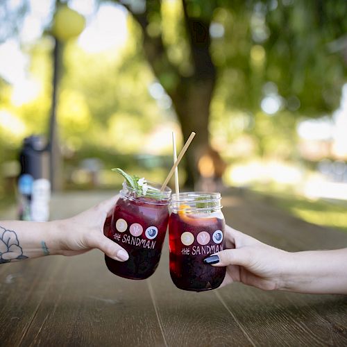 Two people are clinking mason jar glasses filled with a dark-colored beverage outdoors.