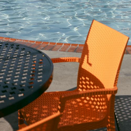 Two orange chairs and a black metal table sit by the edge of a tranquil pool, casting geometric shadows on the ground.