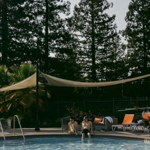 Two people sit by a pool, with sun loungers and shade sails. Trees line the background, adding a peaceful atmosphere to the scene.