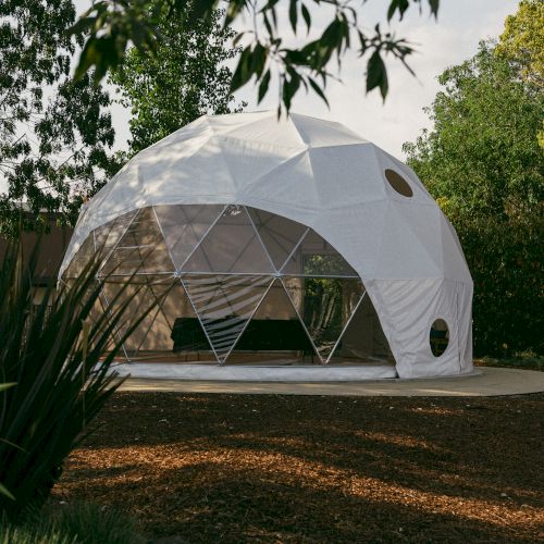 A geodesic dome with a white cover stands surrounded by trees and plants on a circular base in a garden setting.